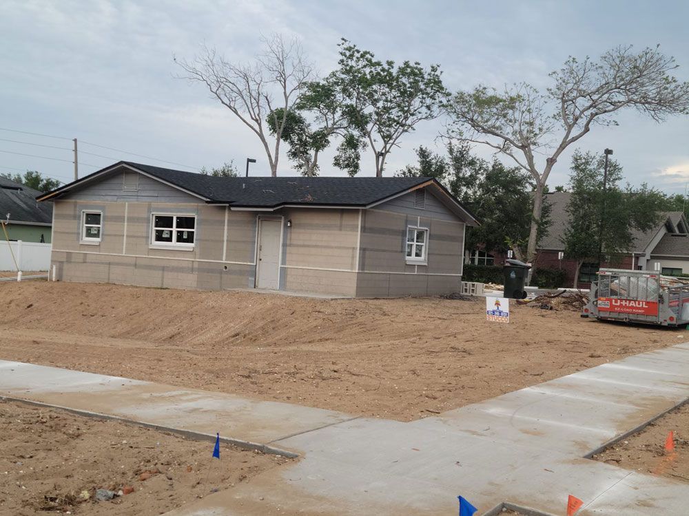 A partially constructed one-story house on a dirt lot with concrete sidewalks. Gray siding and black roof.