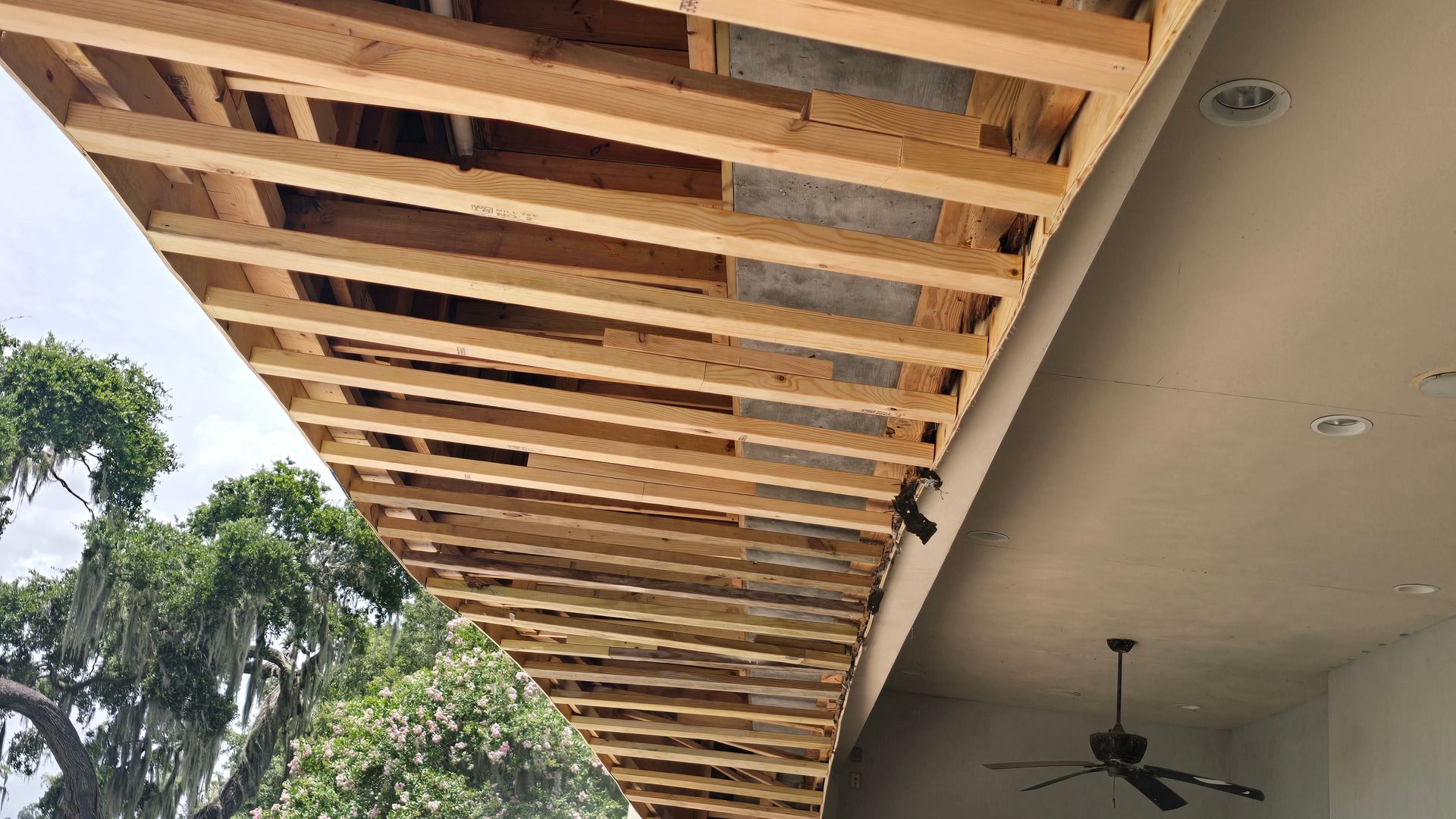 Wooden beams form an open ceiling under an overhang; a ceiling fan hangs on the right. Trees are visible on the left.