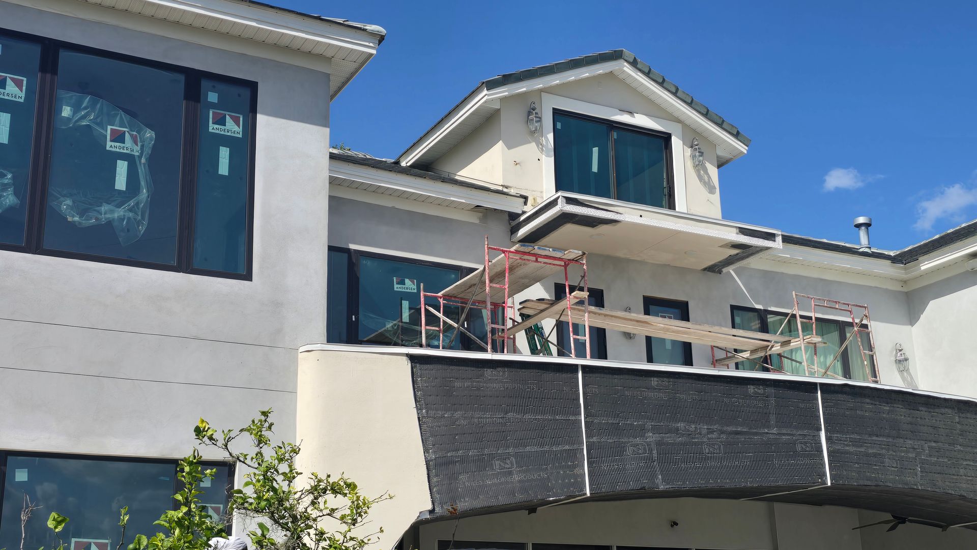 A two-story modern house under construction. Scaffolding sits on the balcony, windows are covered, and the sky is blue.