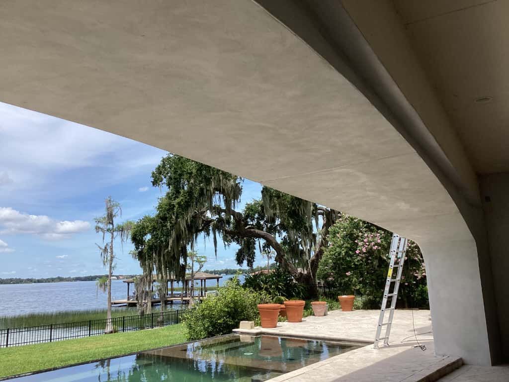 View of a lake and trees through a curved archway. A pool and lawn are in the foreground; a ladder leans against a wall.