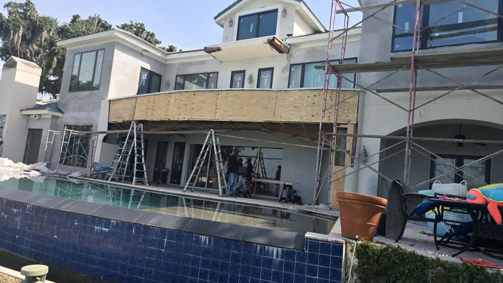 Exterior view of a house under construction with a swimming pool. Scaffolding, exposed wood, and workers are visible.