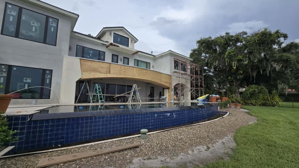 A large, two-story house under construction with a blue-tiled pool in the foreground and a cloudy sky overhead.