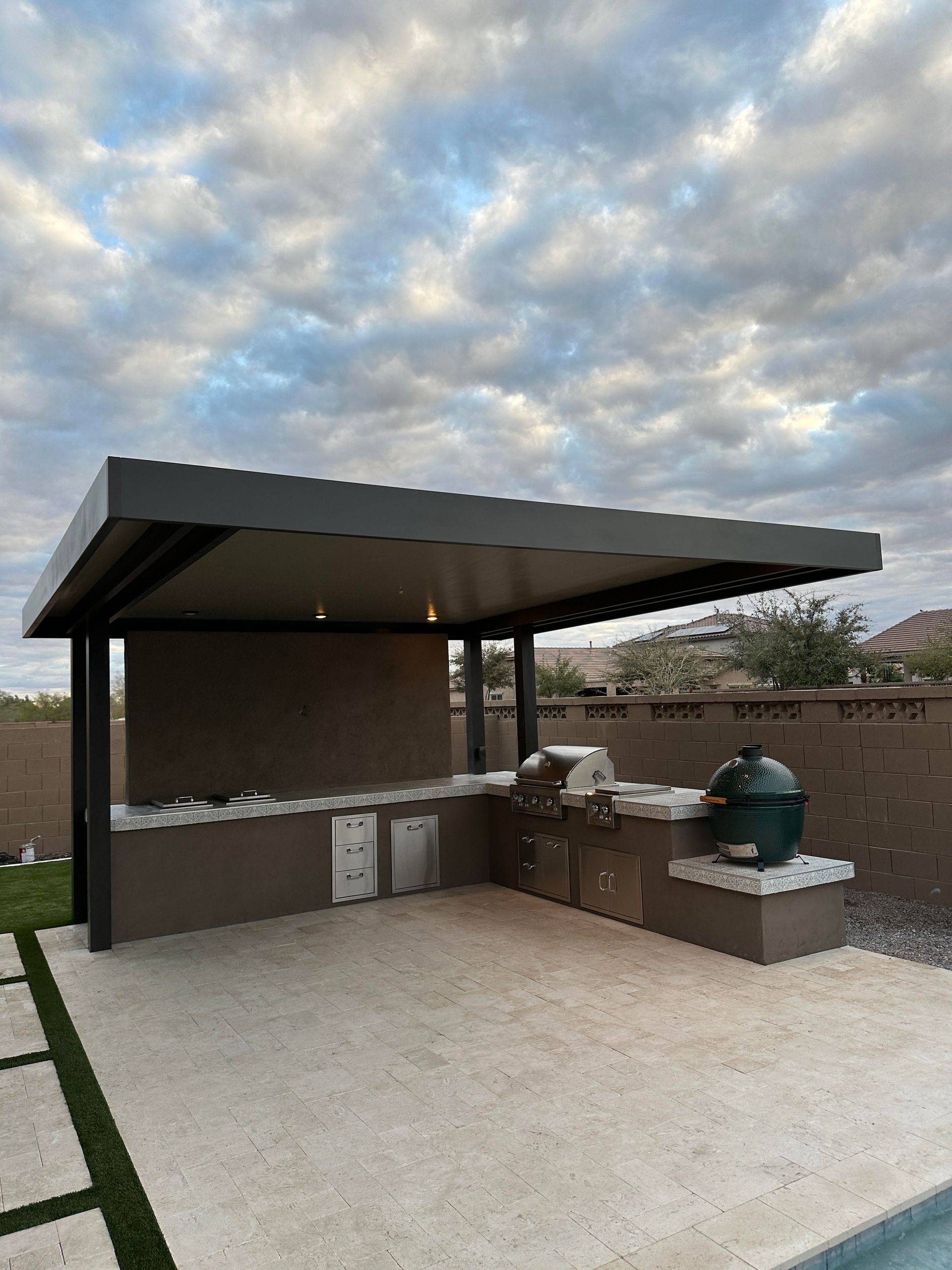 A large green egg grill is sitting under a covered patio next to a swimming pool.