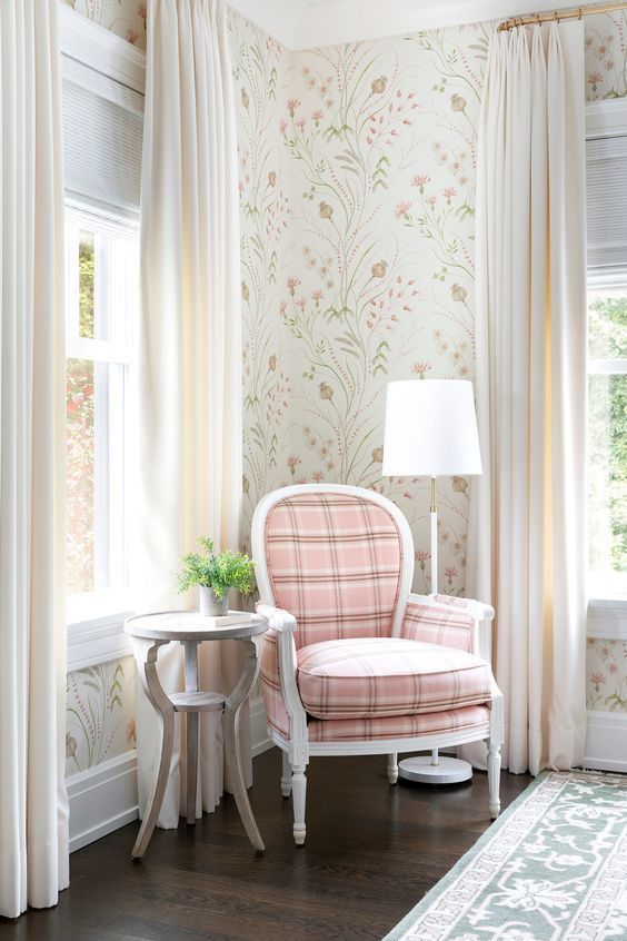 Cozy reading nook with a pink plaid chair, side table, and floral wallpaper, framed by white curtains and windows.