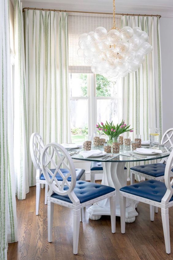 Dining room with round glass table, white chairs with blue cushions, and a bubble chandelier.