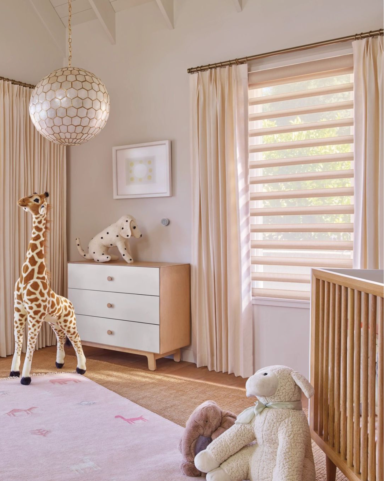 Child's room with stuffed giraffe and sheep, a white dresser, and light-colored curtains and blinds.