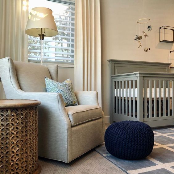 A nursery with a gray crib, beige armchair, and a blue ottoman on a light blue rug.