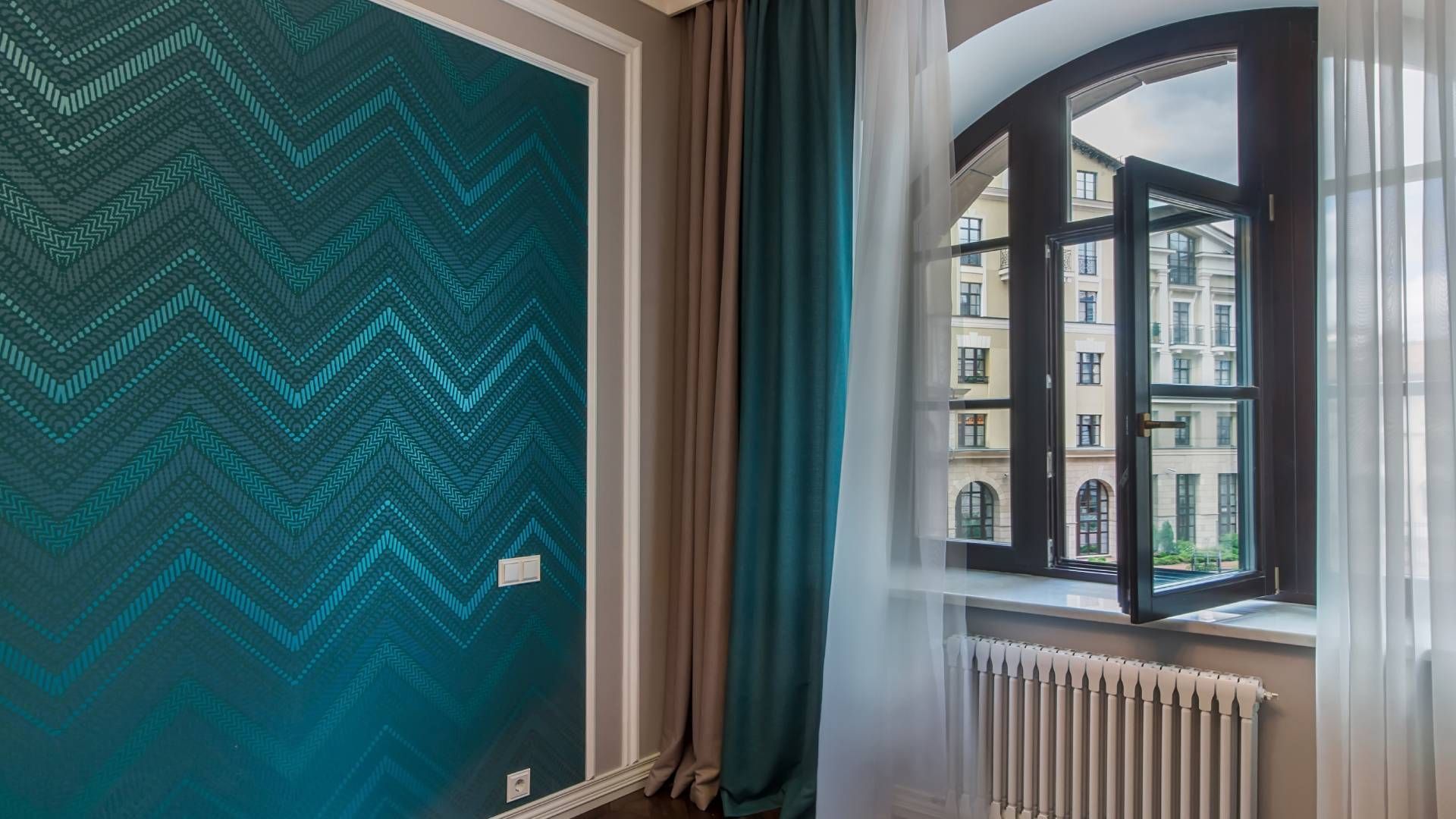Teal zigzag wallpaper next to a window with buildings in view; beige and teal curtains; a radiator.