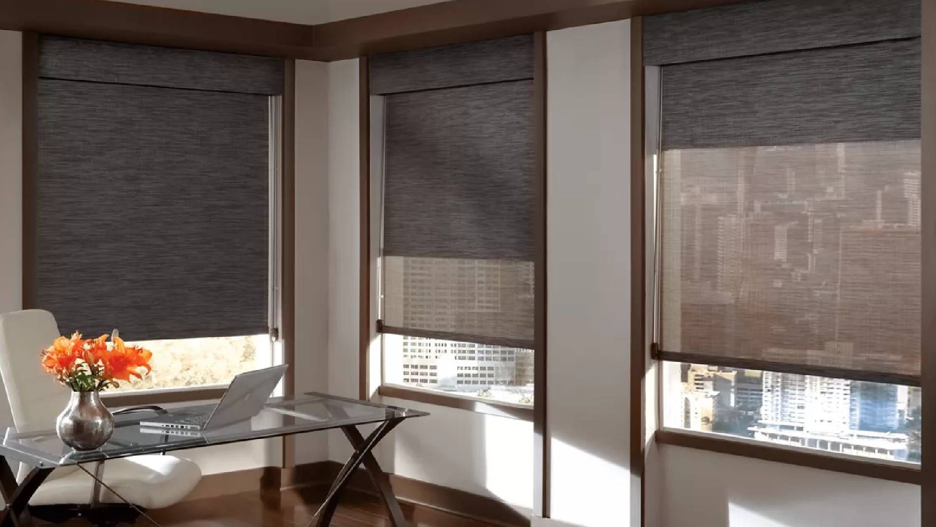 Three windows with dark roller shades, partially open, overlooking a cityscape. Office setting with desk, chair, and flowers.