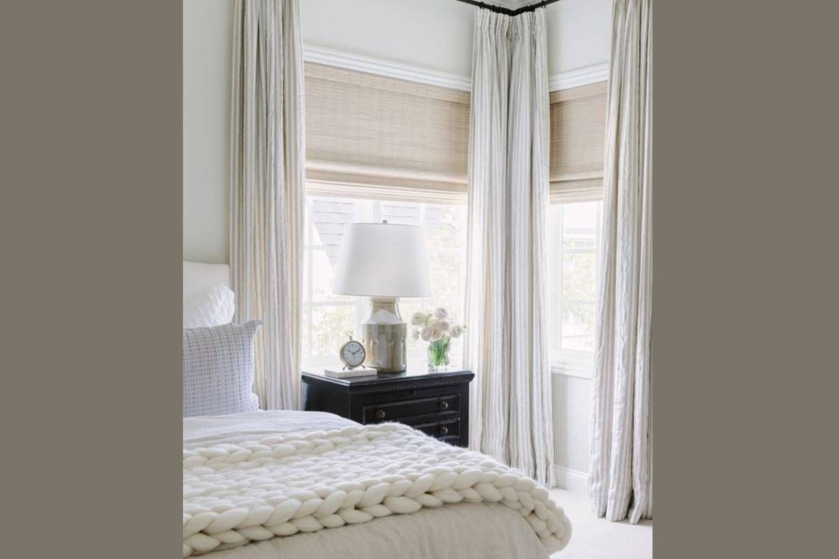 Bedroom with draped curtains, woven shades, bedside table, lamp, and chunky knit blanket.