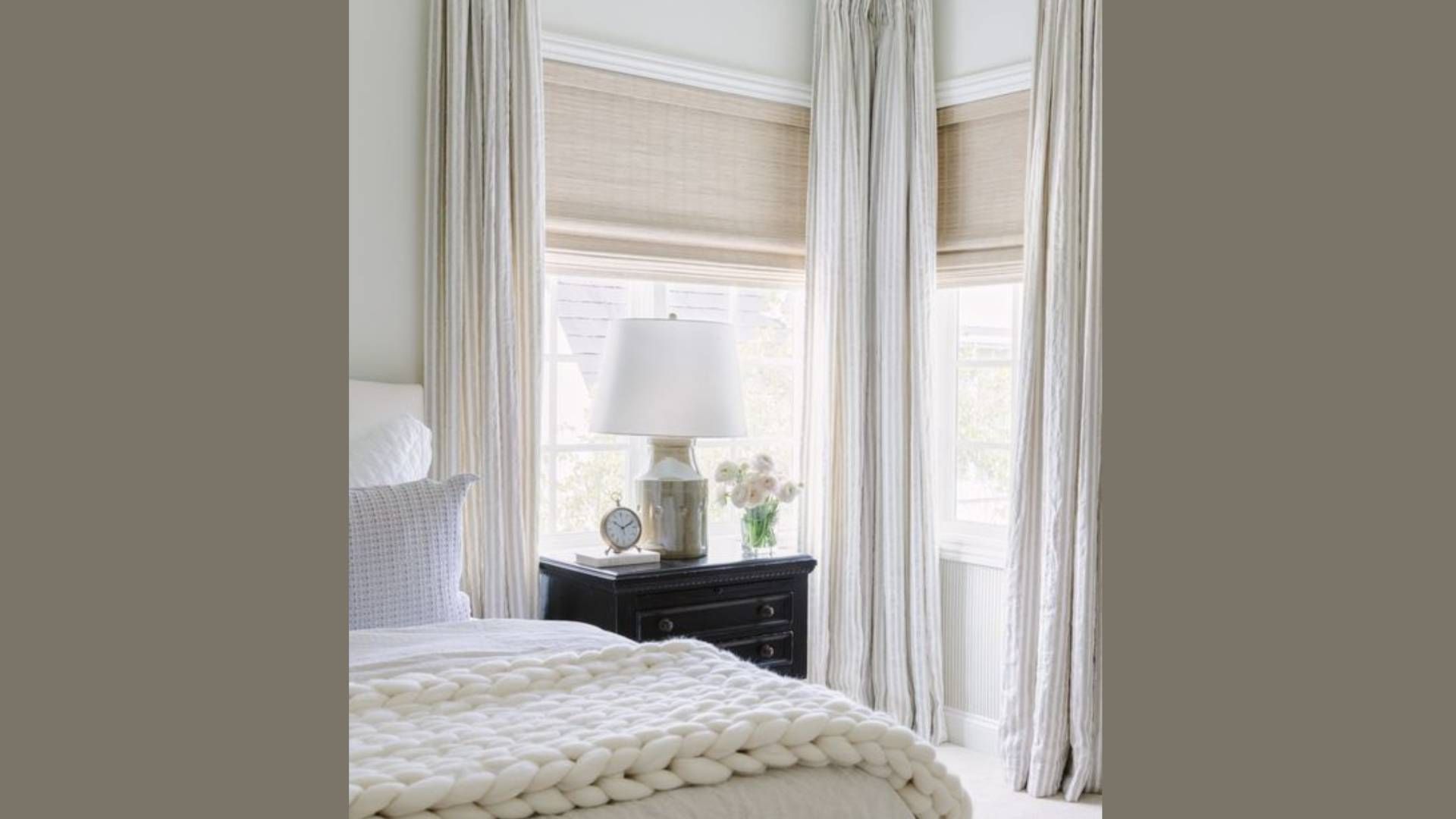 Bedroom with white curtains and woven shades, nightstand with lamp and flowers.