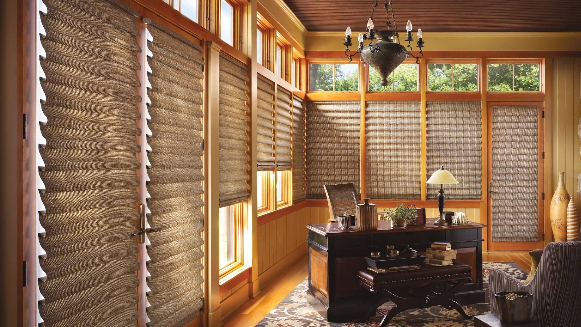 Office interior with wood-framed windows and textured window shades; desk, chair, and chandelier.
