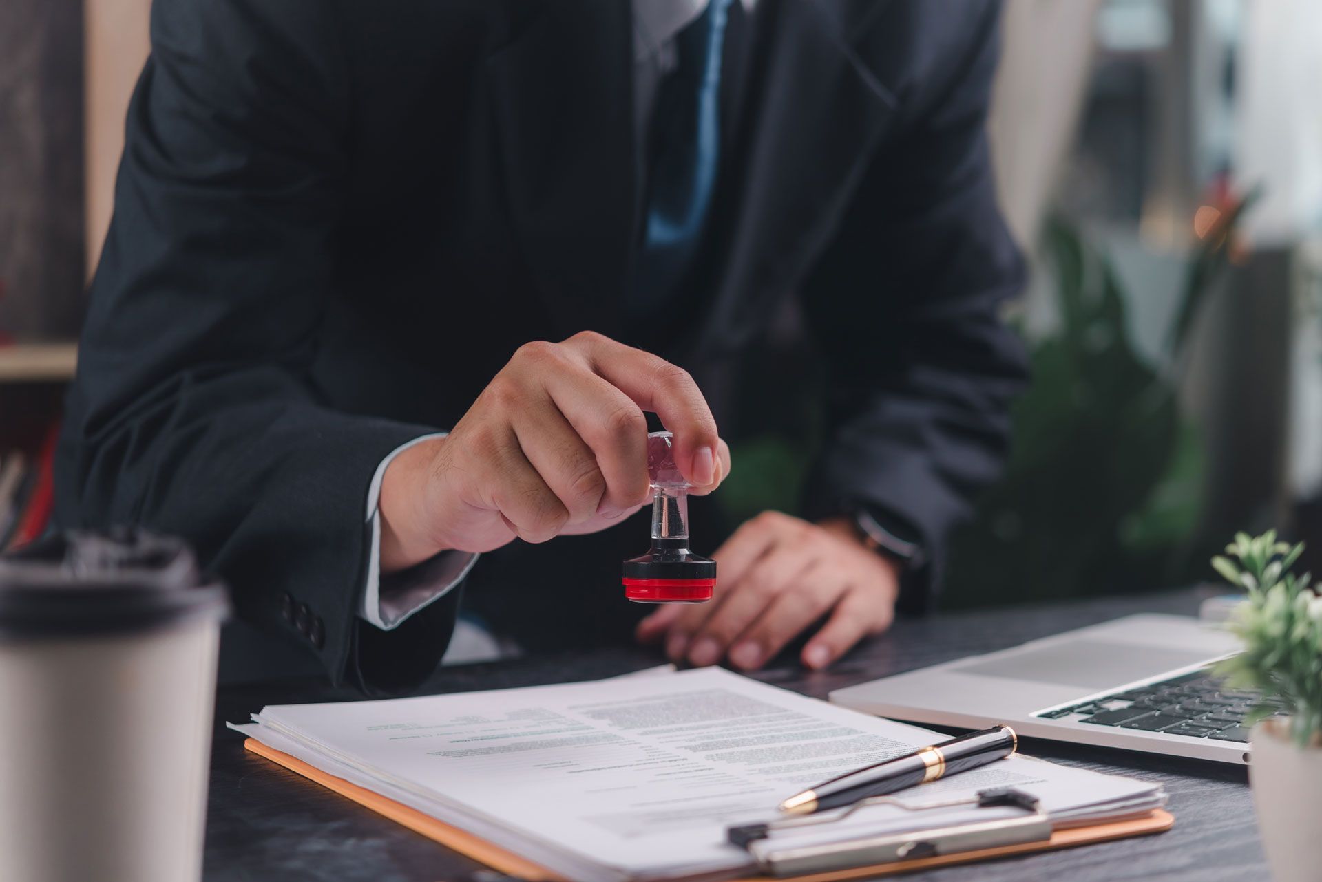 Person in suit stamping a document at a desk with a laptop, pen, and coffee.