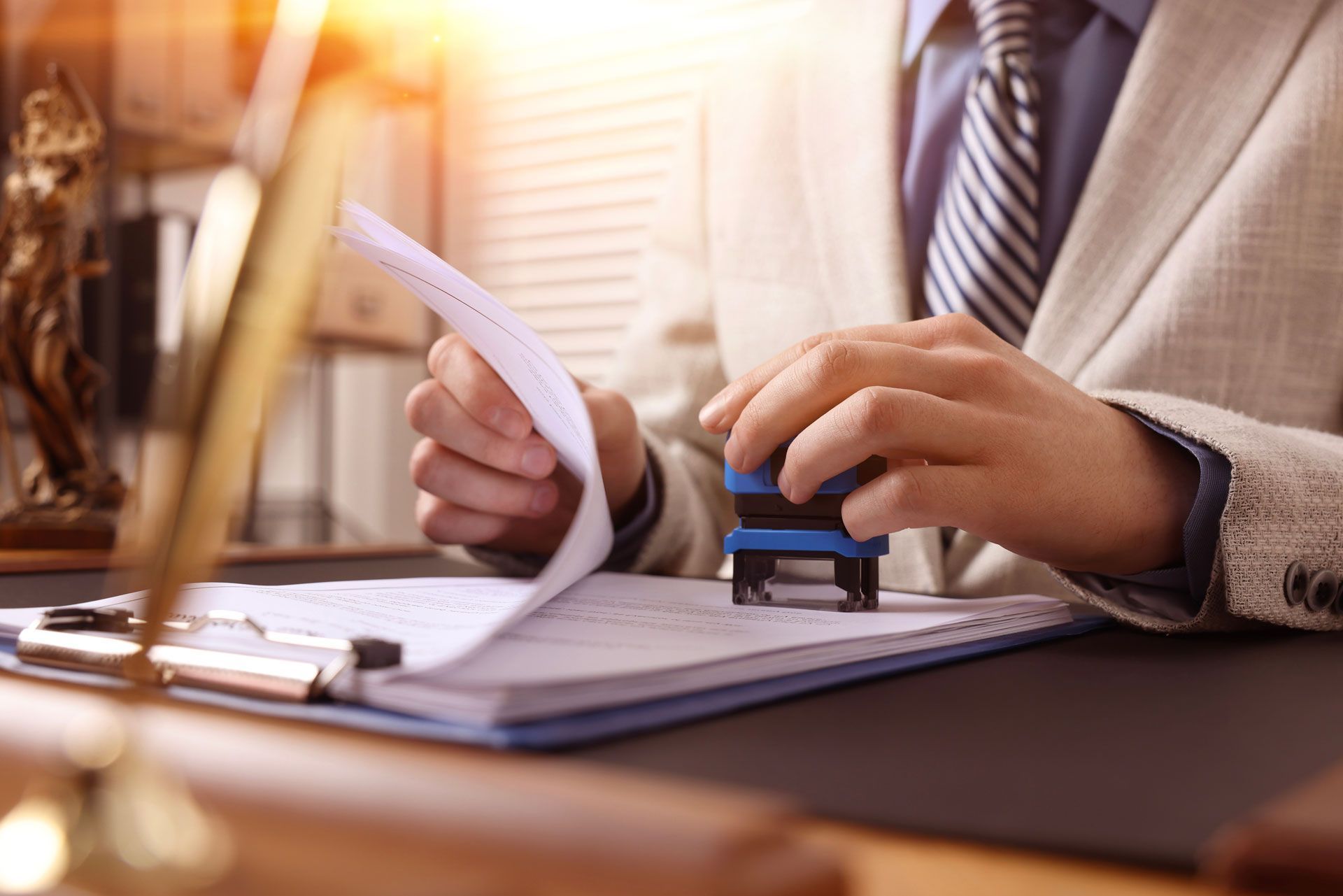 Person in suit stamping a document on a clipboard. Desk with a golden gavel and law book in the background.