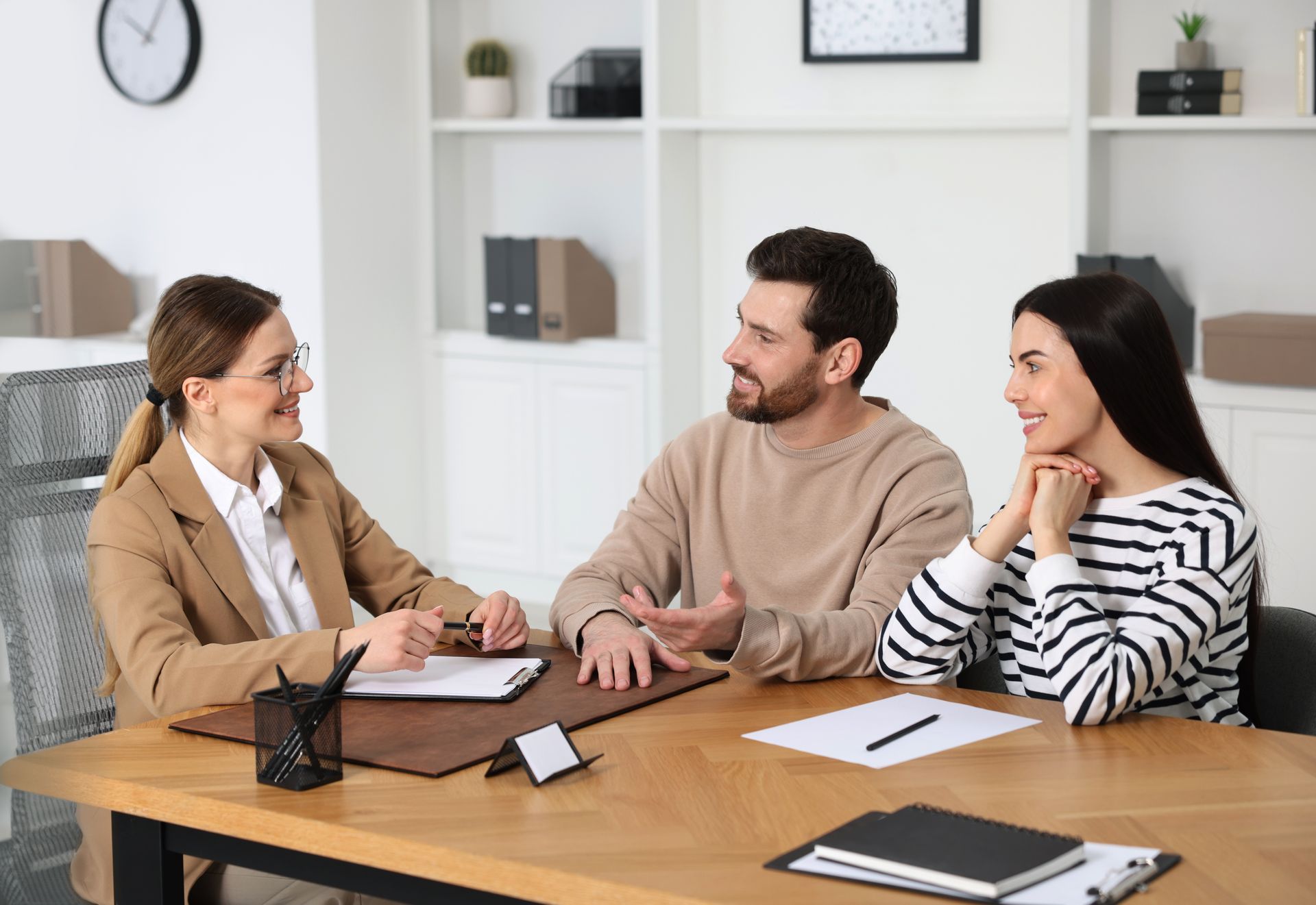 A professional woman in a blazer consults with a couple at a desk in an office setting.