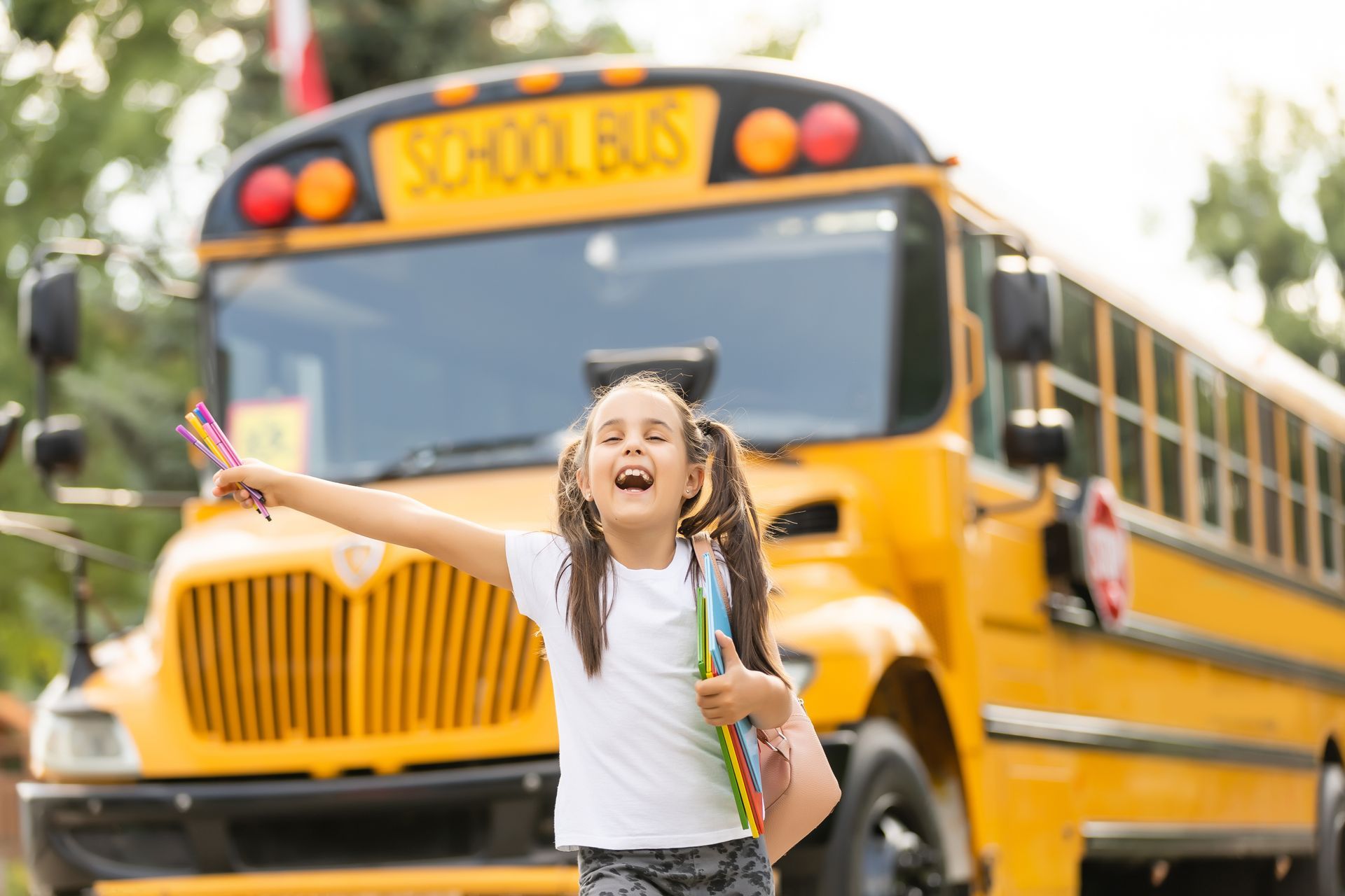 Girl with pigtails laughing, holding books and colored pencils, in front of a yellow school bus.