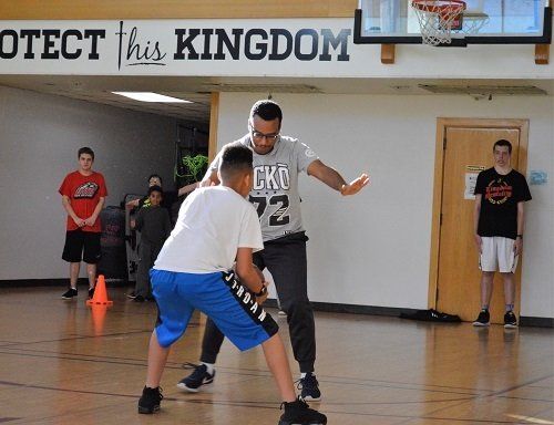 Two boys are playing basketball in a gym with a sign that says protect this kingdom.