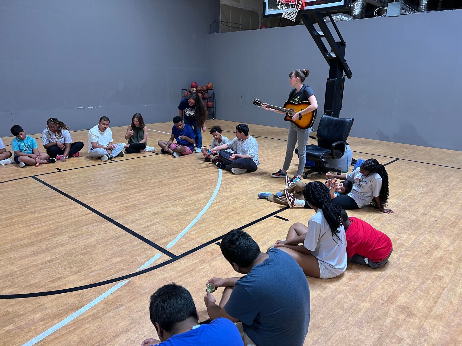 A group of people are sitting on the floor of a basketball court while a woman plays a guitar.