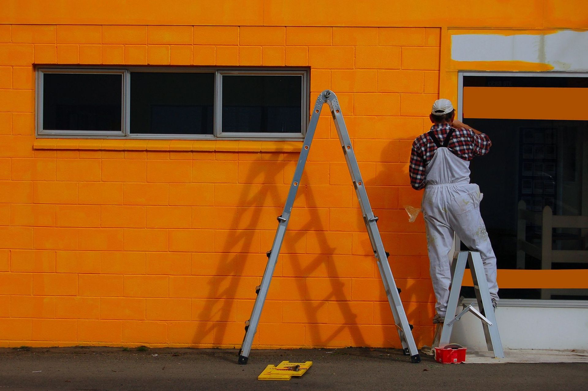 A man is standing on a ladder painting an orange building.