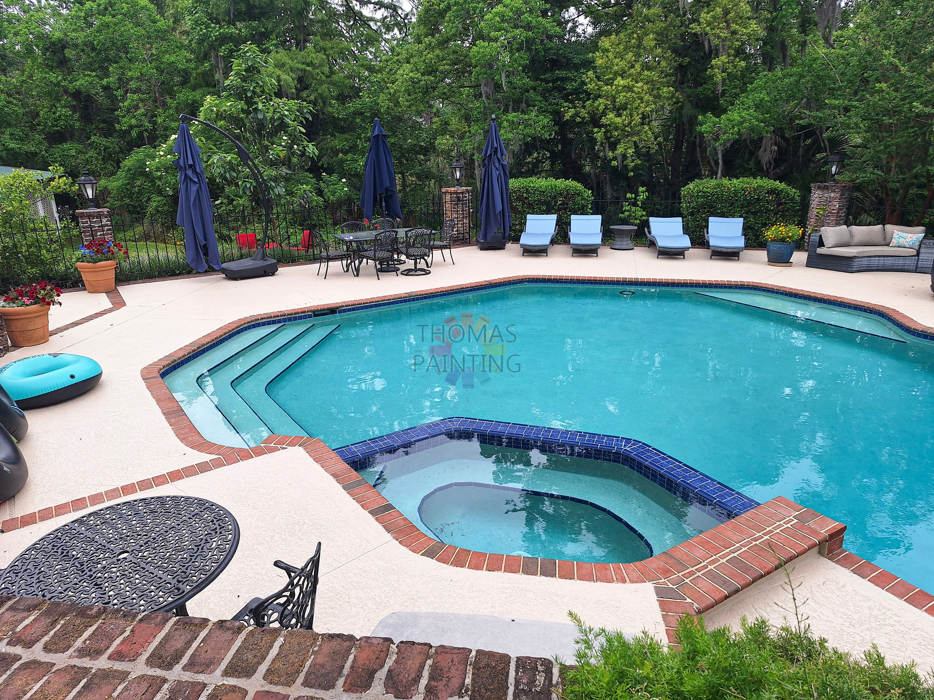 Backyard pool with hot tub, blue umbrellas, lounge chairs, and potted plants amid trees.