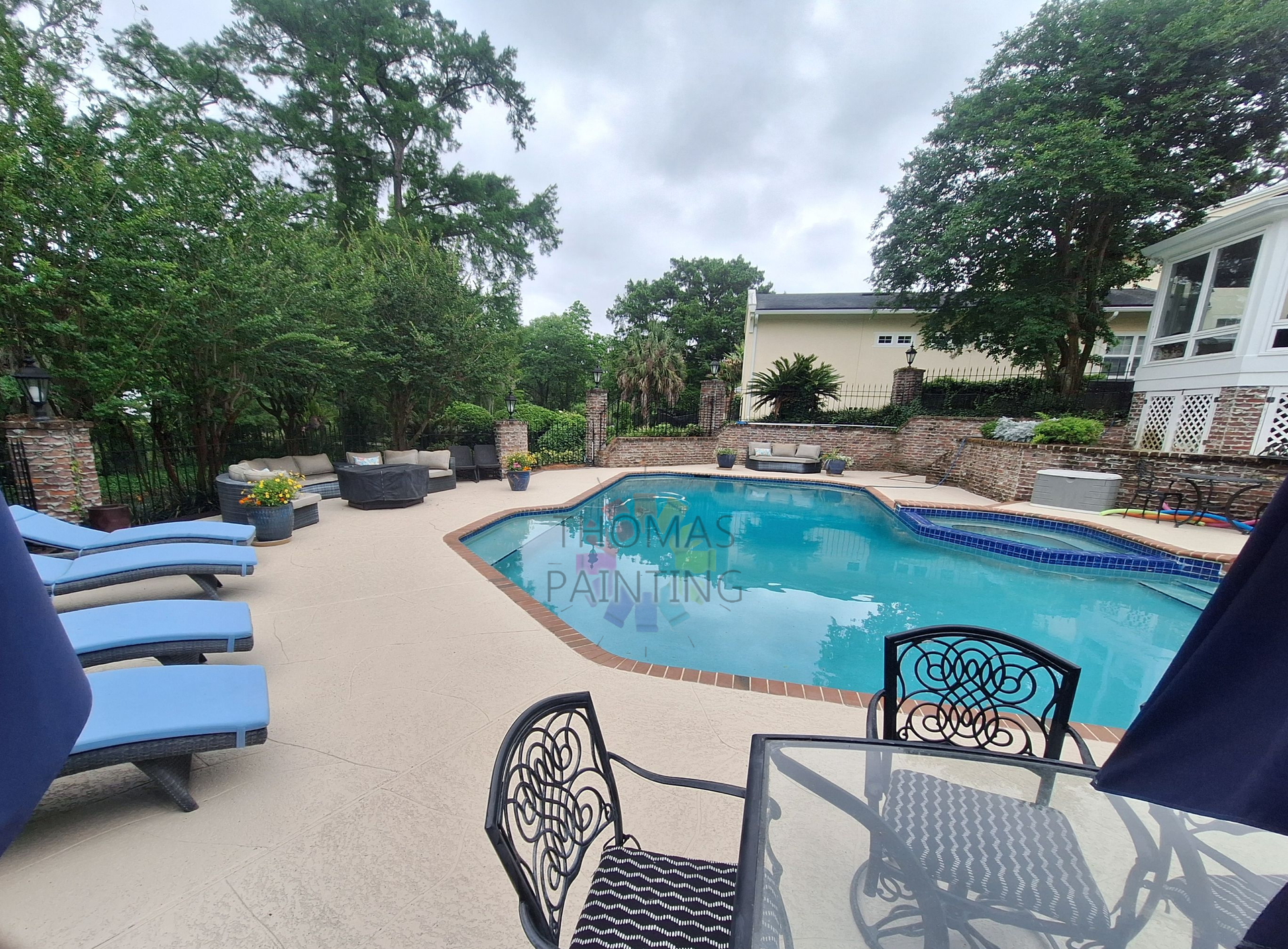 Poolside patio with blue lounge chairs, glass table, and a curved swimming pool surrounded by trees.