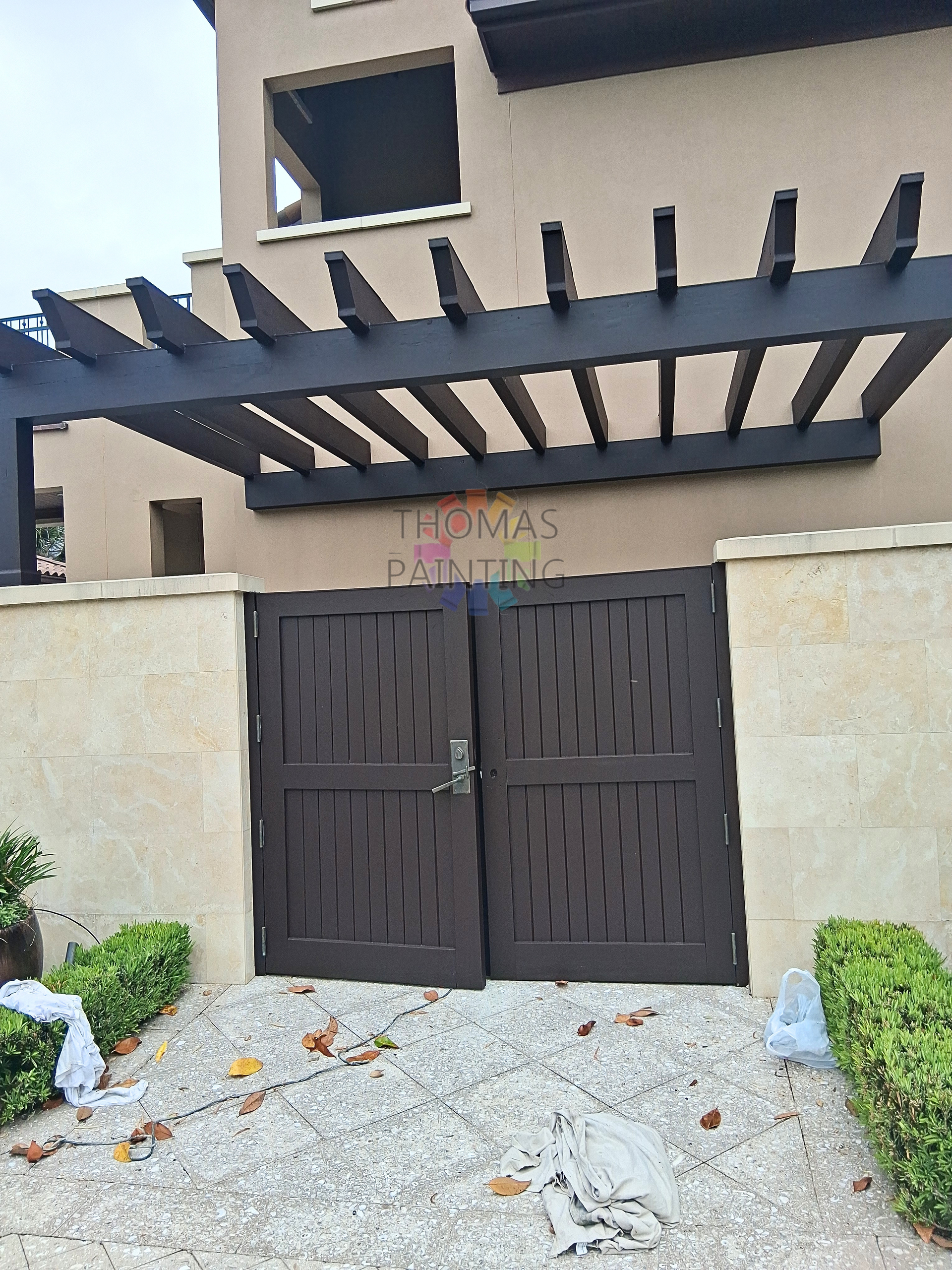 Dark double gate beneath a modern pergola at a beige townhouse entrance with gravel path and shrubs