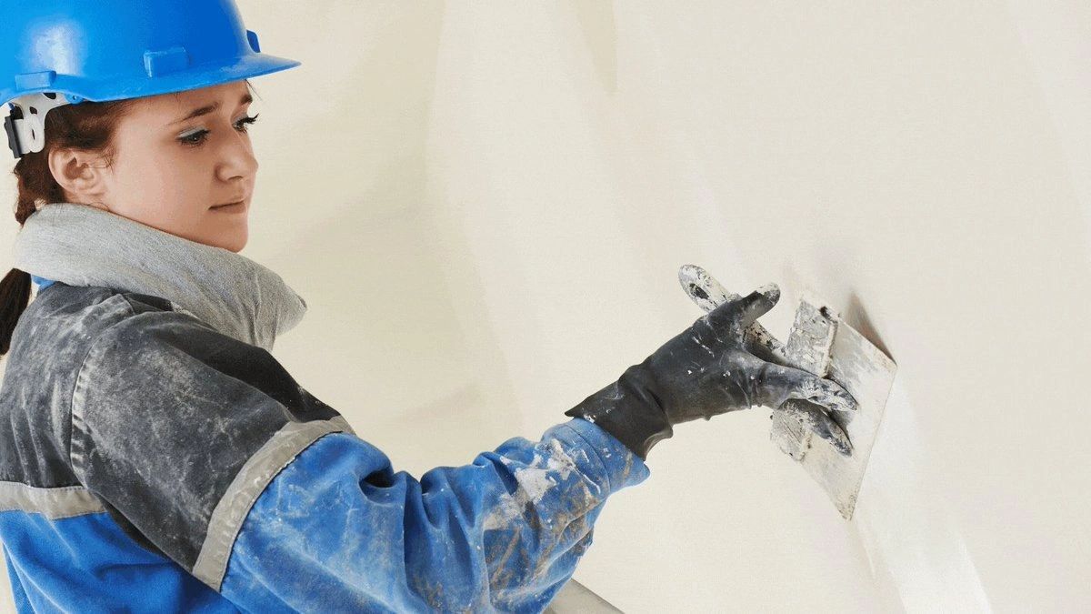 A woman wearing a hard hat and gloves is plastering a wall.