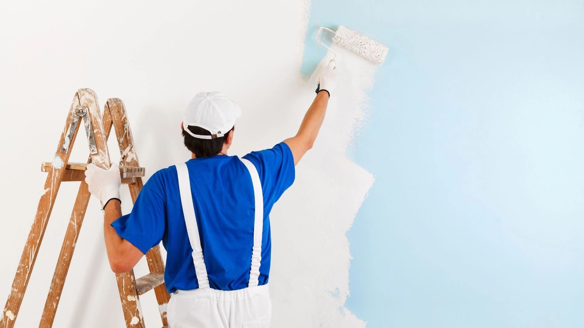 A man is painting a wall with a roller while standing on a ladder.