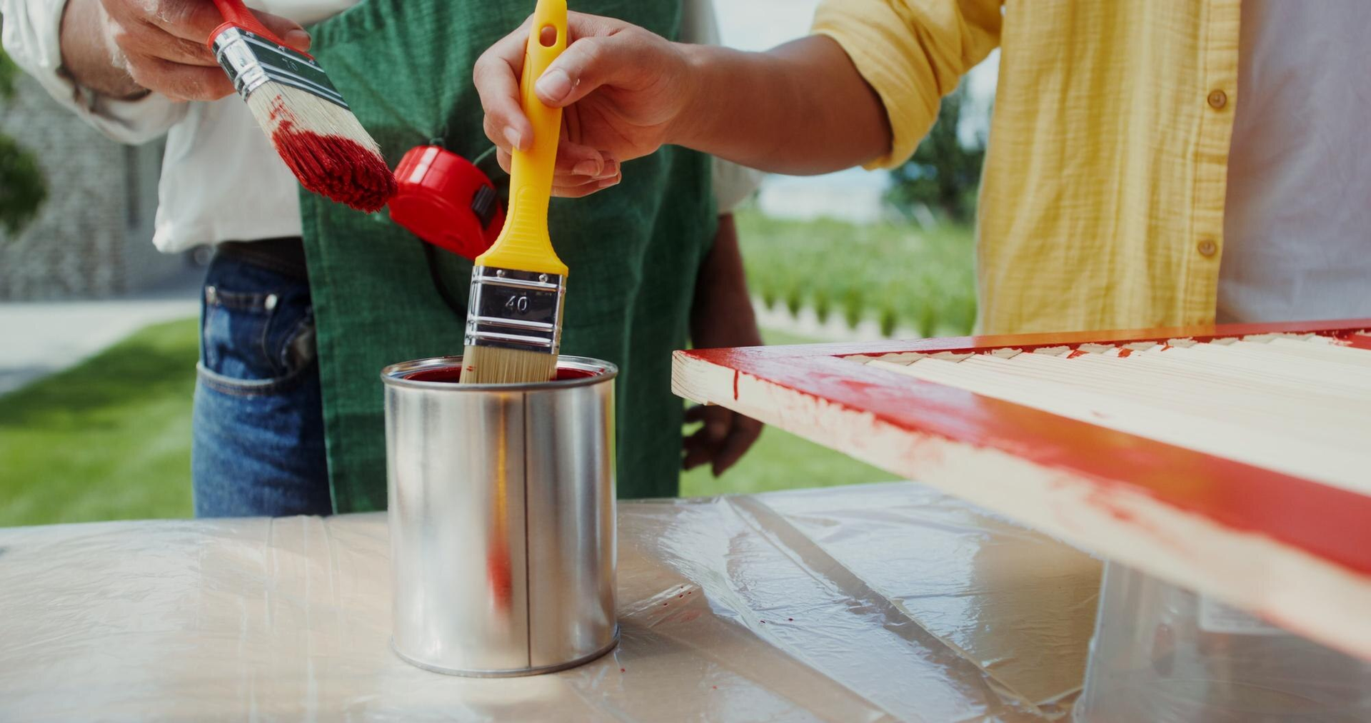 People painting a red-and-white surface with brushes and a paint can outdoors