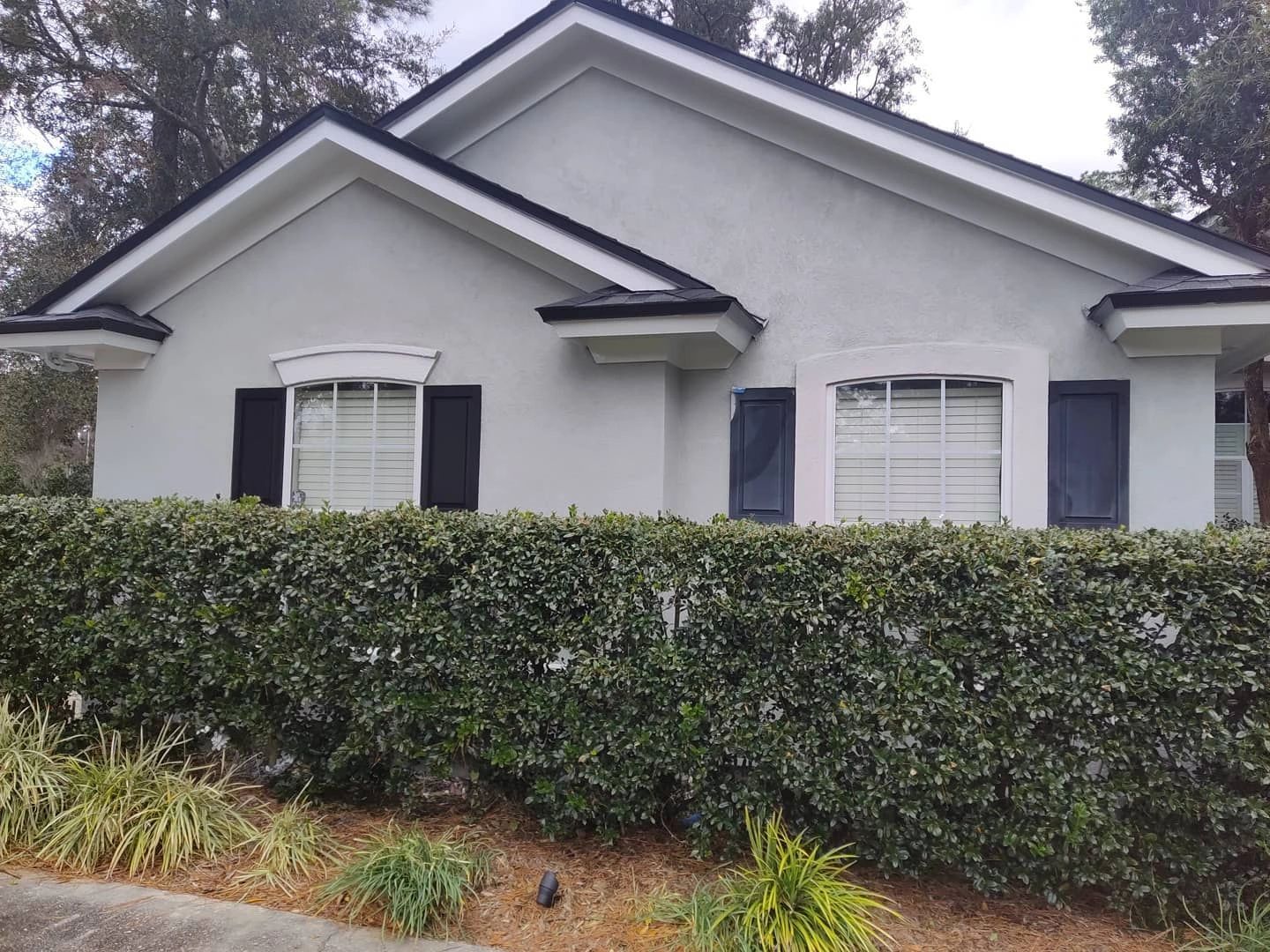 A white house with black shutters and a hedge in front of it