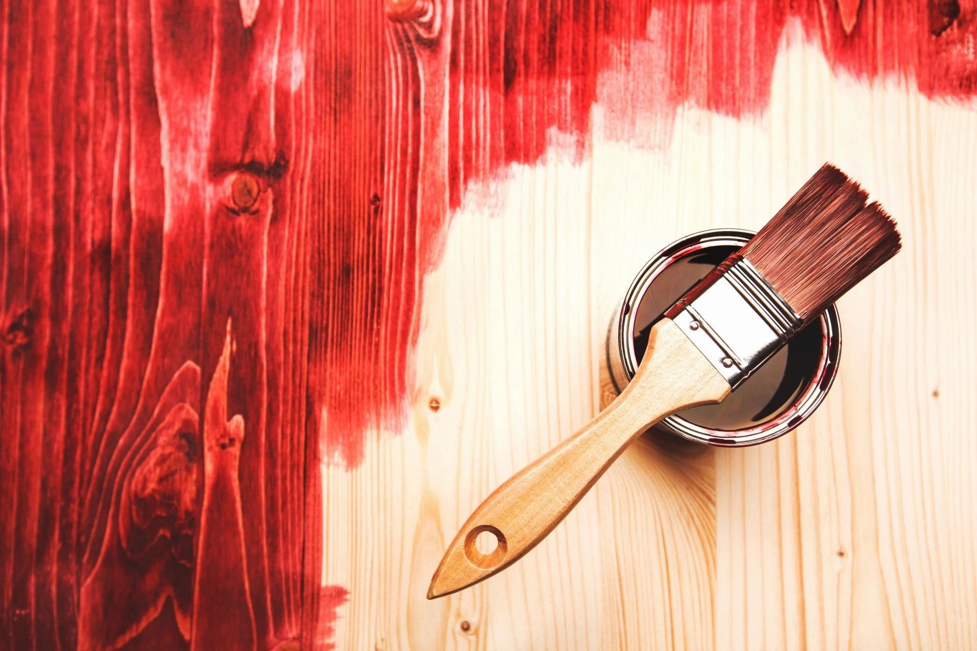 A brush is sitting in a can of red paint on a wooden table.