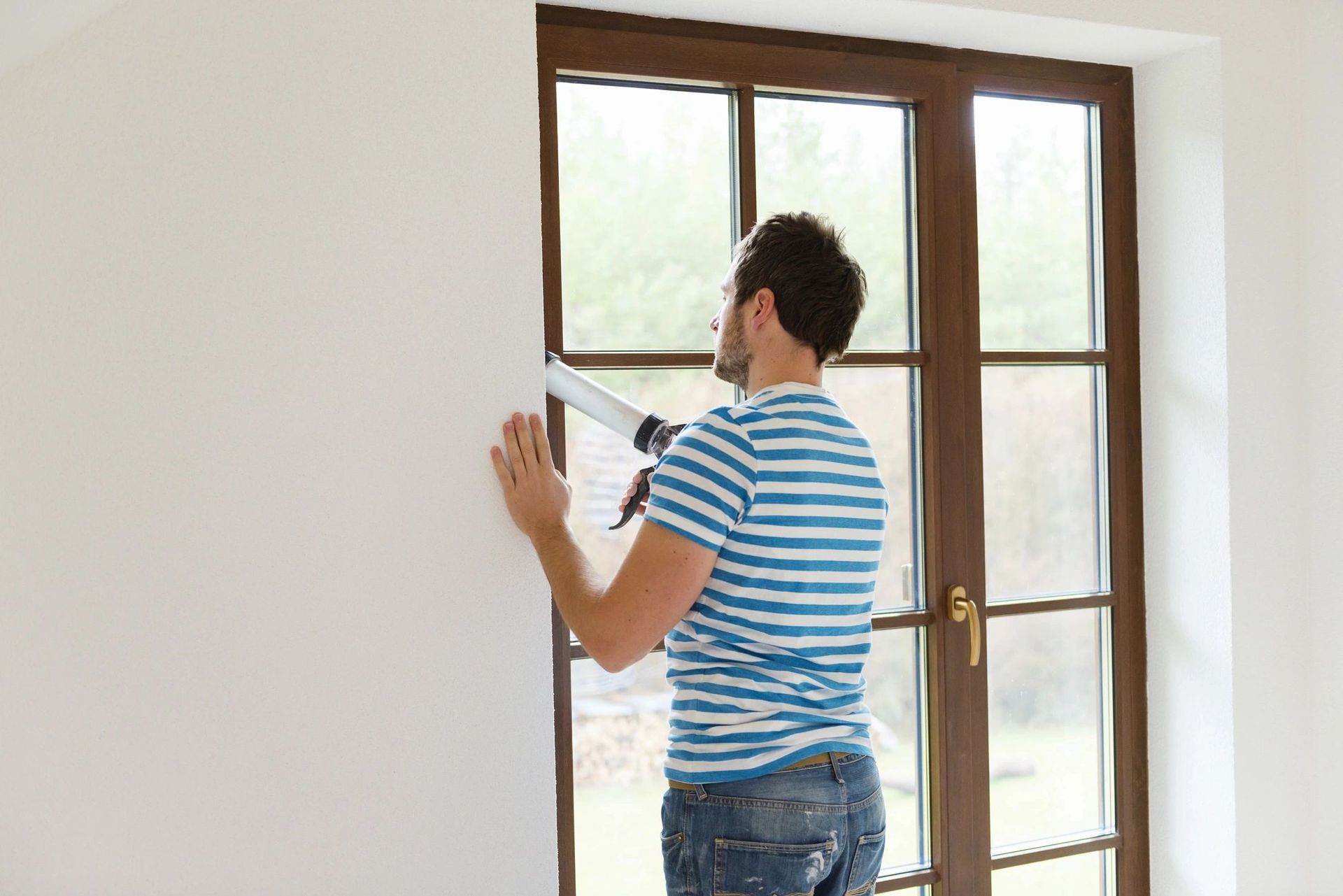 A man is applying caulk to a window in a room.