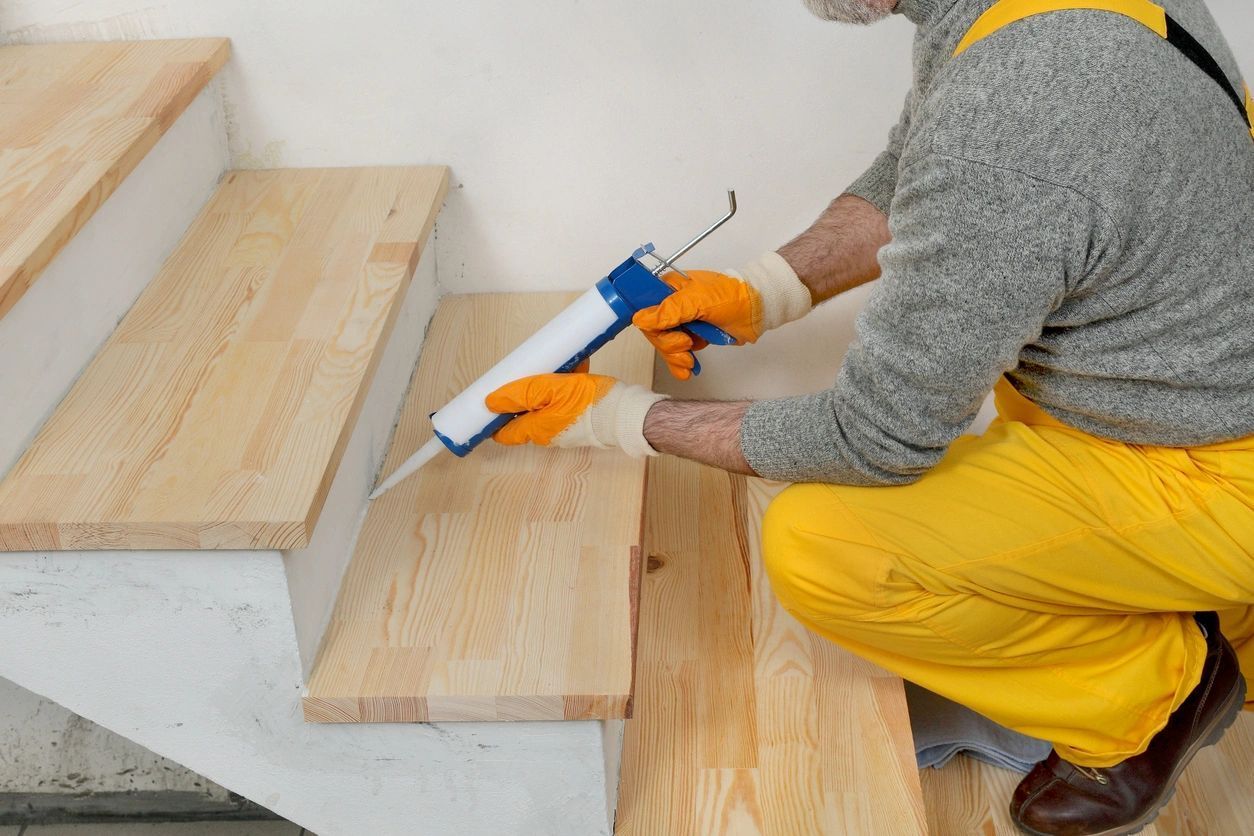 A man in yellow pants is kneeling down and applying glue to a wooden staircase.