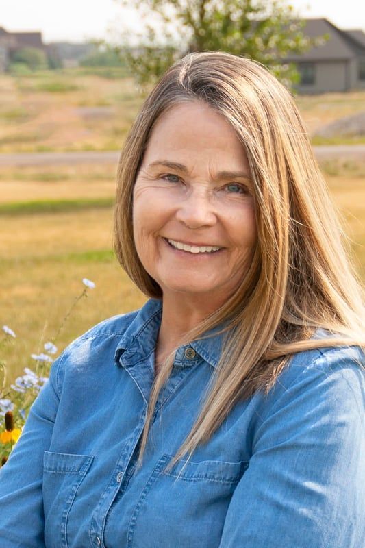 Woman with long blonde hair smiles, wearing a blue denim shirt, outdoors with yellow field backdrop.