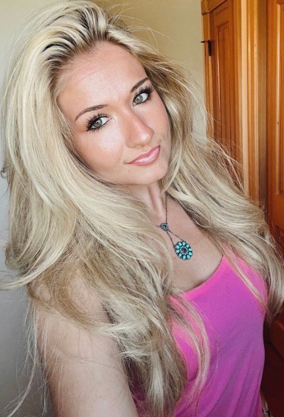 Blonde woman with long hair wearing a pink top, smiling, turquoise pendant.