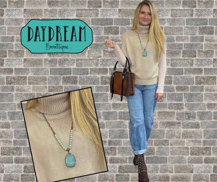 Woman in beige sweater, blue jeans, boots, and turquoise necklace, holding a brown purse. Daydream Boutique logo in background.