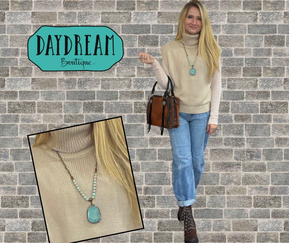 Woman in jeans, sweater, and necklace, holding a bag, posed in front of a brick wall, Daydream Boutique logo.