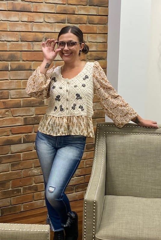 Woman in glasses, jeans, and floral top, leaning on chair in front of a brick wall.