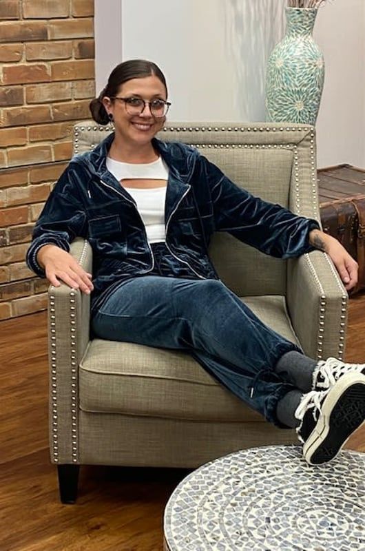 Woman sitting in armchair, wearing blue velvet jacket and pants, white top, glasses, and sneakers.
