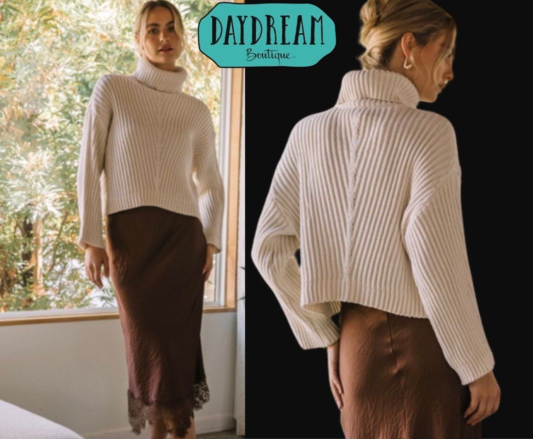 Woman wearing a cream turtleneck sweater and brown satin skirt; poses in front of a window and against a black background.