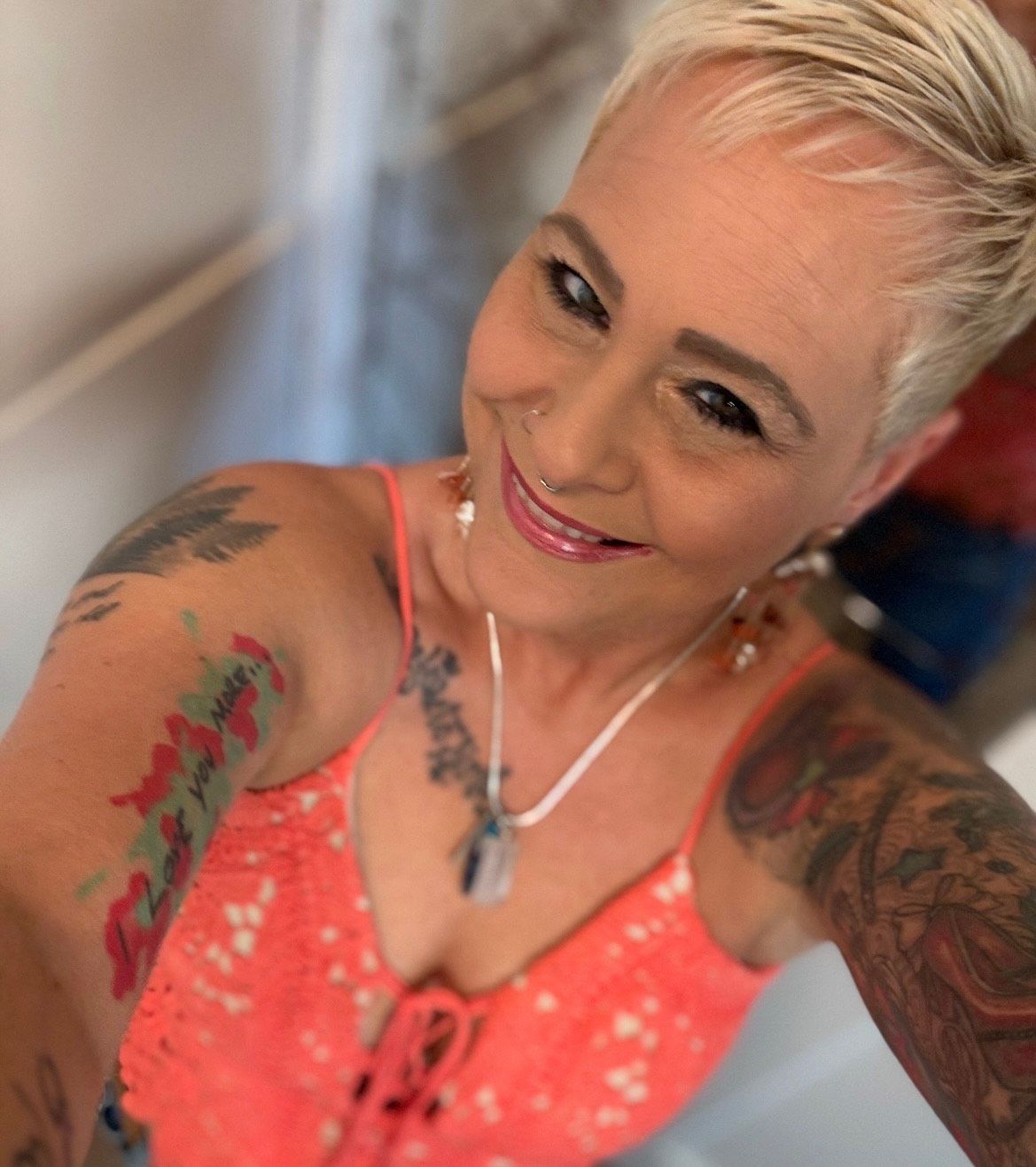 Woman with short blonde hair, tattoos on arms, wearing an orange top, smiling, looking at the camera.