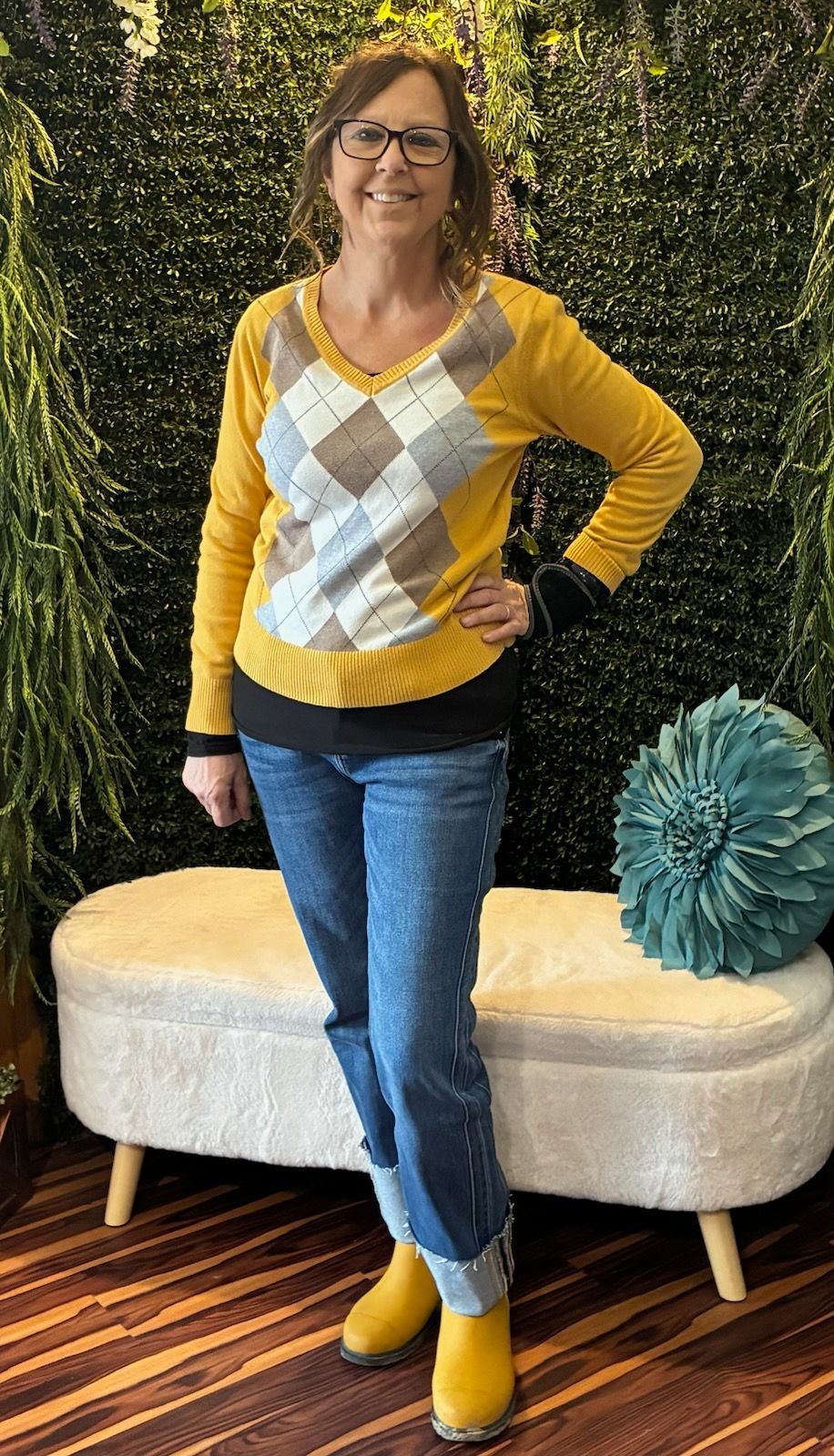 Woman in yellow sweater and blue jeans poses indoors; smiles.