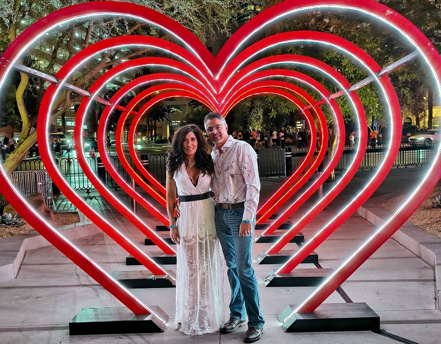 Couple standing in front of a series of red, heart-shaped light structures; night setting.