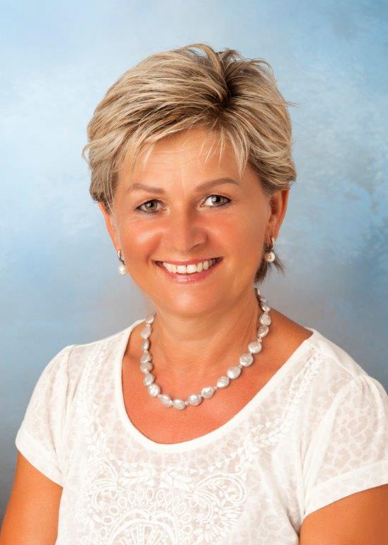 Frau mit kurzen blonden Haaren, lächelnd, trägt ein weißes Hemd und eine Perlenkette.