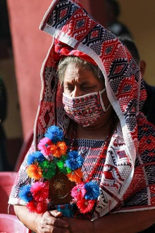 Frau in traditionell rot-weiß gemusterten Textilien, dazu passende Maske. Bunte Blumengirlande um den Hals.