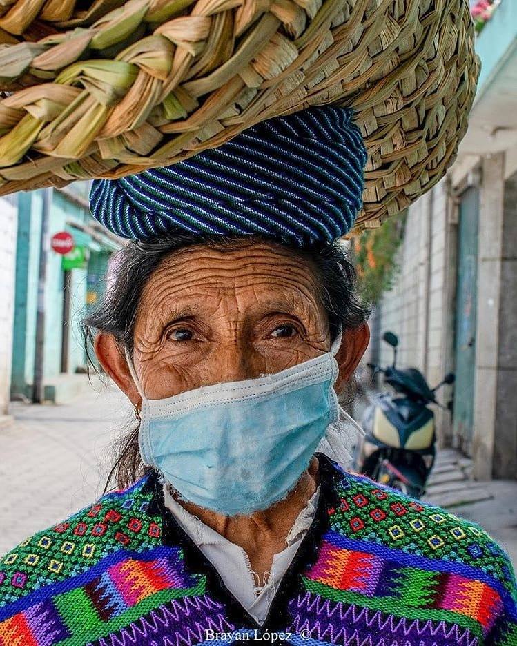 Eine Frau in einem farbenfrohen, bestickten Kleidungsstück, die eine Gesichtsmaske trägt und einen Korb auf dem Kopf hat,