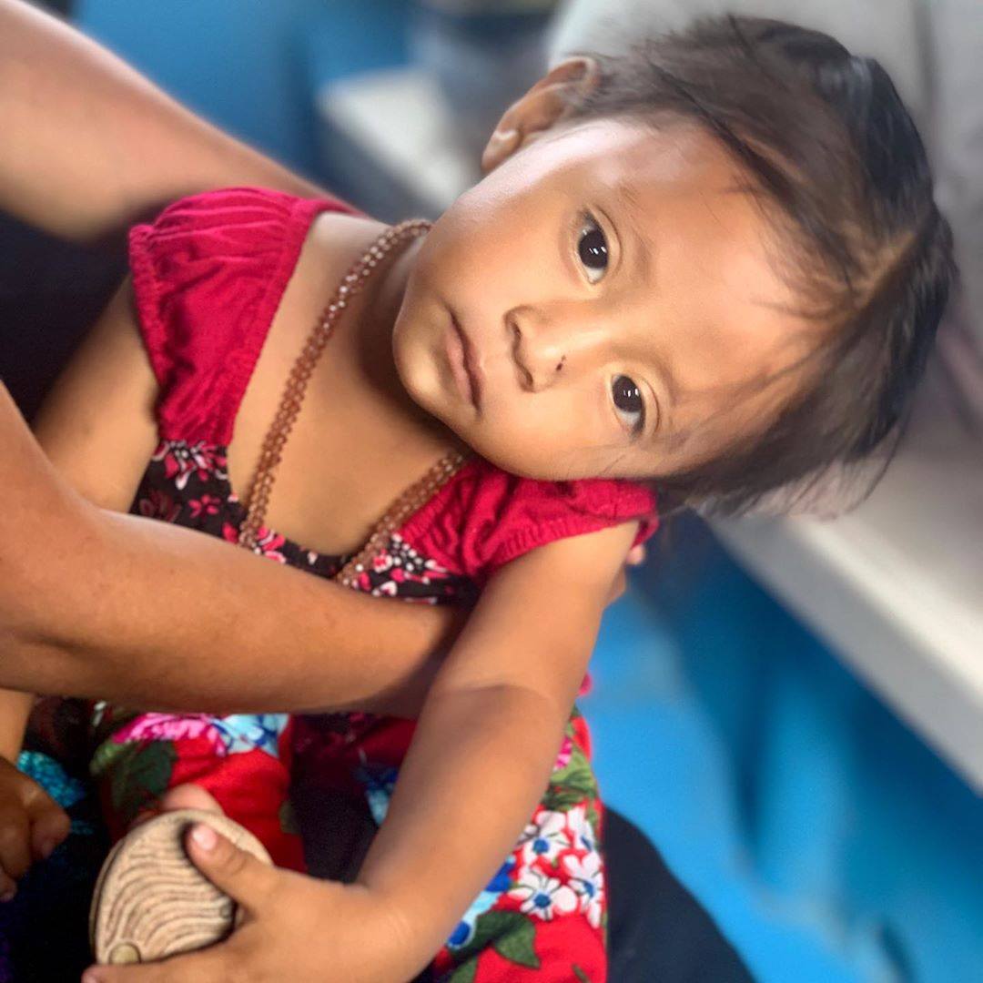 Kind in rotem Blumenkleid, Kopf geneigt, wird im Haus gehalten.