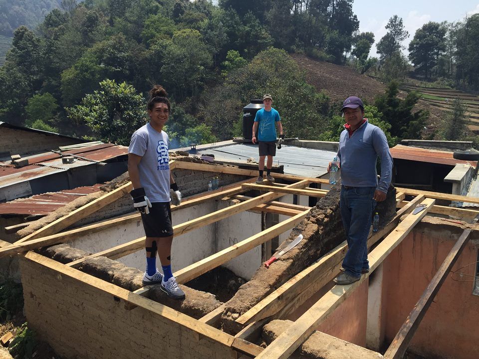 Drei Personen errichten auf einem Dach Holzstützen. Im Hintergrund Berge und Grün.