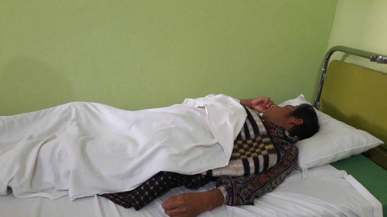 Eine Person liegt im Bett und ist mit einem weißen Laken zugedeckt. Eine grüne Wand und ein Metallrahmen sind zu sehen.