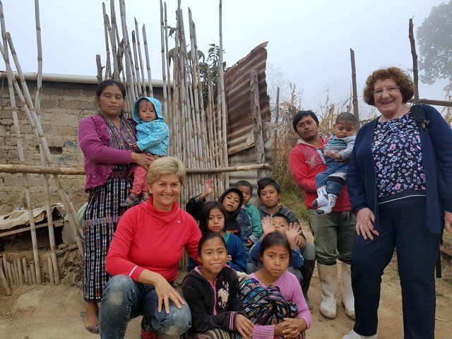 Die Gruppe posiert vor einem rustikalen Gebäude. Erwachsene halten Kinder im Arm; andere sitzen oder stehen lächelnd.